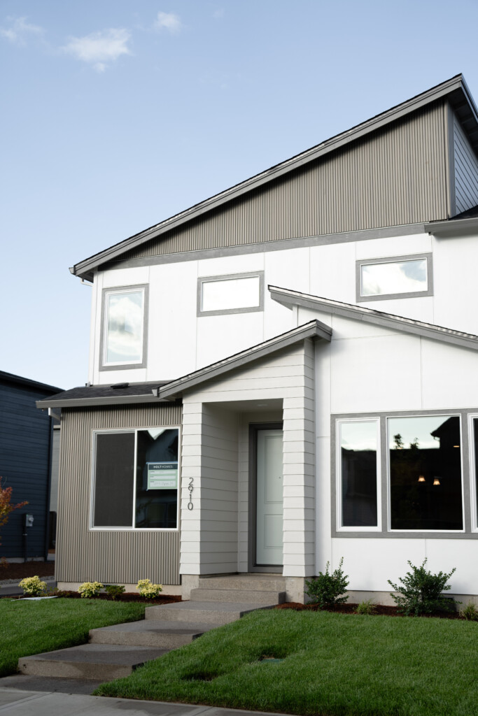 A modern two-story house with gray and white exterior, large windows, and a small covered entryway built through a partnership with West Tuality Habitat for Humanity. Manicured lawn and shrubs line the front beneath a clear blue sky.
