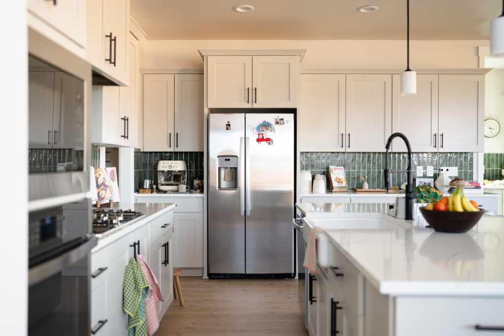 A modern kitchen with white cabinets, stainless steel refrigerator, a kitchen island with a black faucet, fruit bowl, coffee maker, and various kitchen items neatly arranged—ideal for homes with a Distributions Systems Warranty.