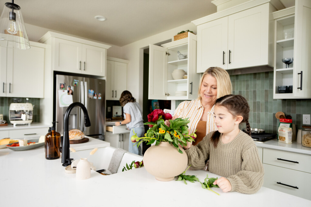 A woman and a young girl arrange flowers in a vase on the kitchen counter, making their new neighborhood feel like home, while another woman works at the sink. The bright kitchen features white cabinets and green tiles.