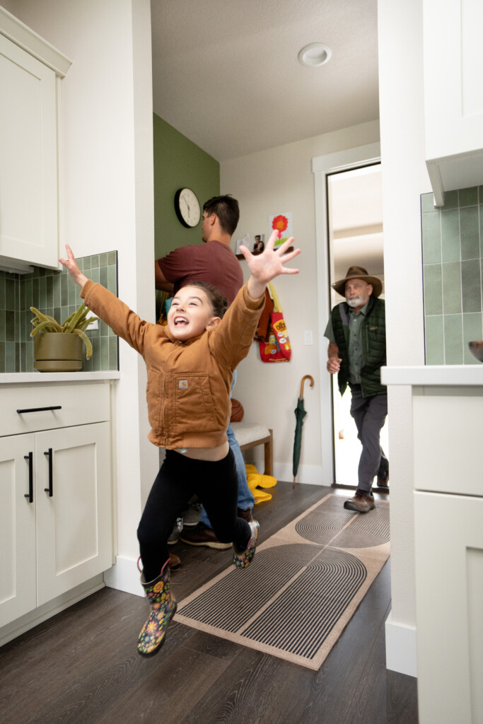 A joyful young girl in a brown jacket and rain boots, ready for water activities, runs with arms outstretched in a bright kitchen as a man hangs up coats and an older man enters the house through the open door.