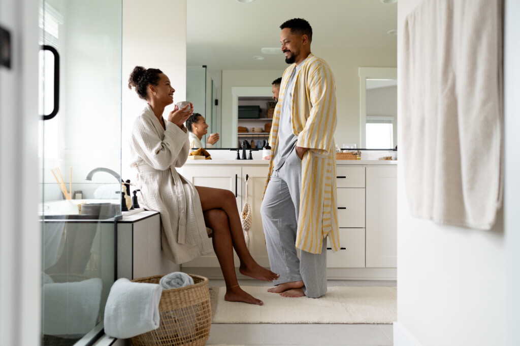 A woman sits on a bathroom counter drinking from a mug while a man stands nearby. Both wear robes and smile at each other in a bright, modern bathroom, enjoying warmth thanks to attentive water heater care, with a basket and towels visible.