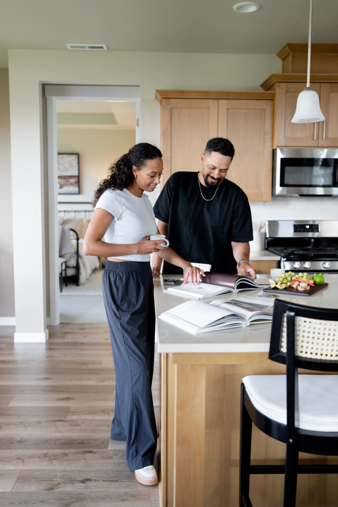 A smiling couple stands in a modern kitchen, looking at open books on the counter as they follow a step-by-step guide. The woman holds a mug, and the man points at a page. Fresh vegetables and a purse are also on the counter.