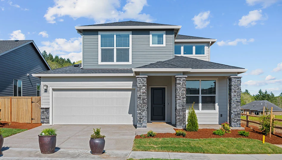 Modern two-story house with gray siding, white trim, and stone accents around the entryway. Located in Ponderosa Ridge, Corvallis Oregon, this new model home features a double garage and neat front yard with small shrubs.