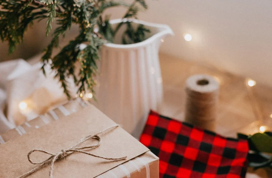 A brown paper-wrapped gift tied with twine sits next to a spool of string, a white ceramic pitcher with greenery, and red-and-black plaid fabric—designer-approved holiday decor with warm fairy lights glowing in the background.