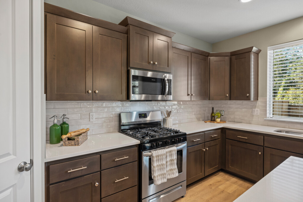 Modern kitchen at 24457 Middlebrook, White Oak Terrace features dark wood cabinets, white countertops, stainless steel gas stove and microwave, white subway tile backsplash, and a window with blinds. Green bottles and utensils accent the counter.