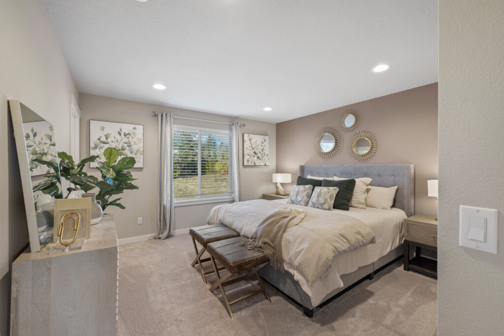 A modern, cozy bedroom with a large bed, neutral bedding, two benches at the foot, nightstands with lamps, a dresser with decor, wall art, and a window with light gray curtains letting in natural light at 24457 SW White Oak Terrace.