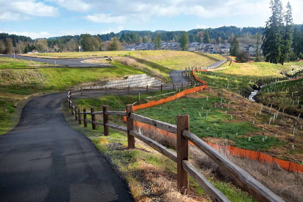 A paved path curves through a grassy landscape in Pleasant Valley Villages, with a wooden fence on one side and an orange construction barrier on the other, leading toward trees and distant houses under a partly cloudy Happy Valley sky.
