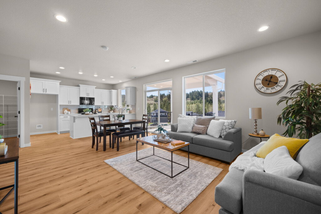 Open-concept living and dining area at NW Tanoak Dr in Ponderosa Ridge features light wood floors, modern gray sofas, a rug, wall clock, indoor plant, and dining table; large windows fill the space with natural light and tree views.