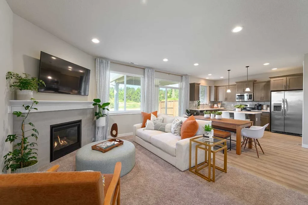 Modern open-concept living room and kitchen, like those in Dream Homes, with light wood floors, a white sofa with orange pillows, fireplace, mounted TV, plants, and large windows. Kitchen features stainless steel appliances.