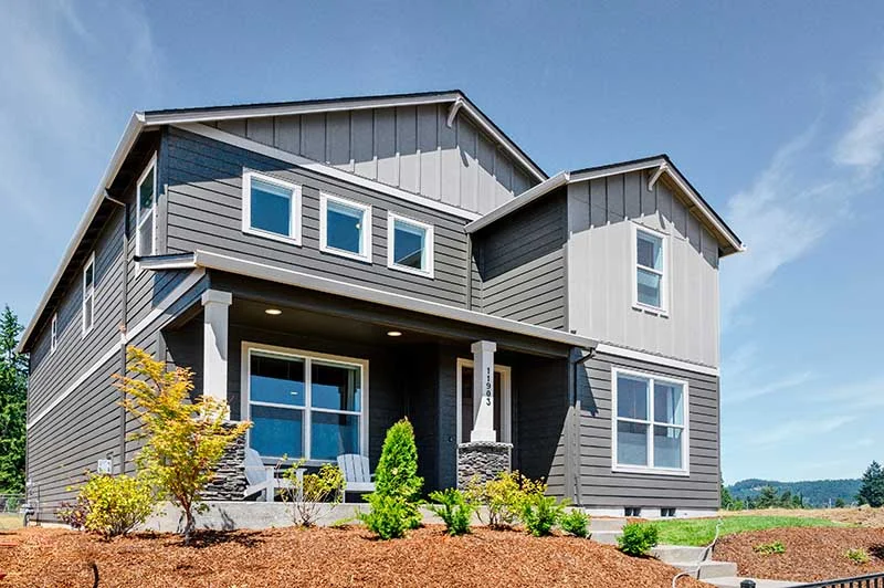 Modern two-story house with gray siding, large windows, and a covered front porch with chairs. Located among the stunning Homes on the Street of Dreams, it features a landscaped yard with shrubs and fresh mulch under a blue sky.
