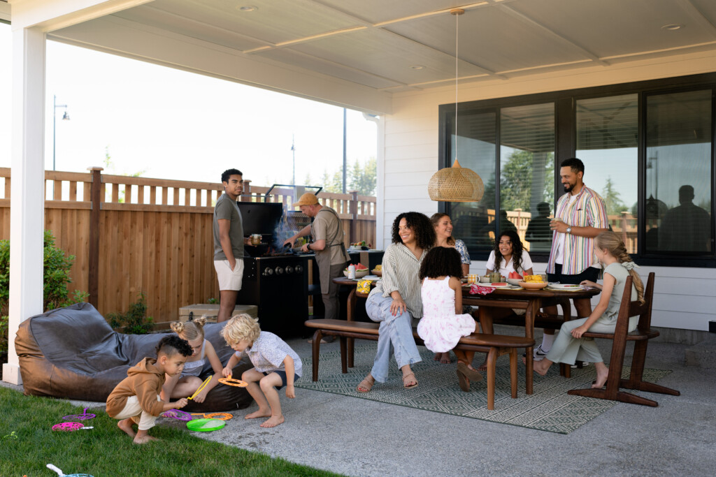 A group of adults and children enjoy a backyard gathering in cozy outdoors. Some adults sit at a table with food, while others grill or stand nearby. Children play on the grass, all under the relaxed shelter of covered outdoor living spaces.