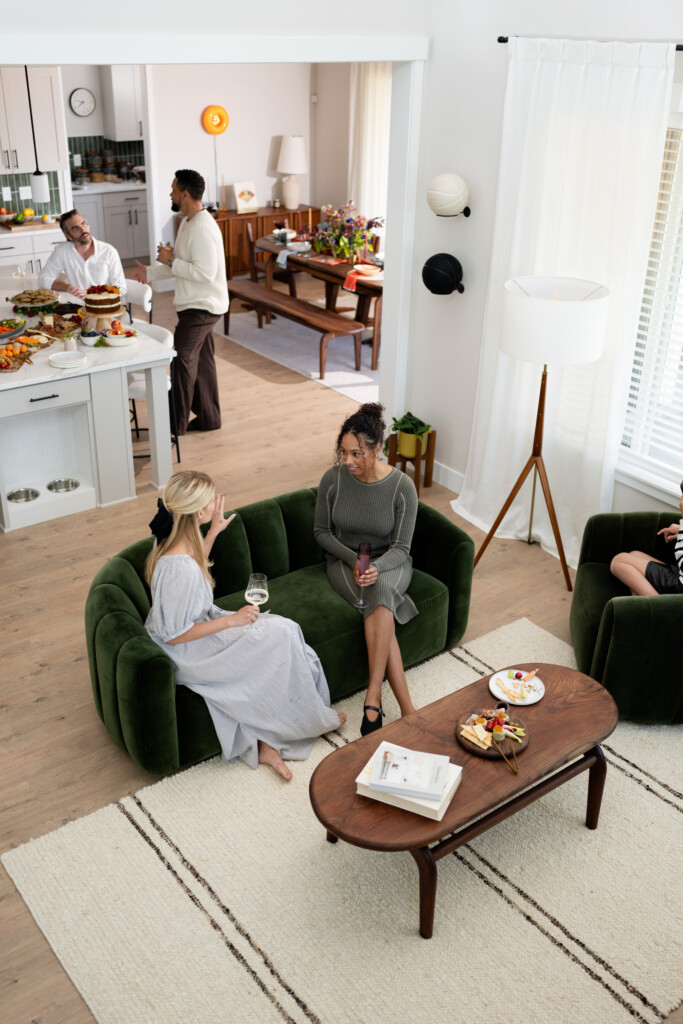 A group of people socialize in a bright living room and kitchen, embodying modern nesting. Two women chat on a green couch with drinks, while others gather near the kitchen island with food and a dining table in the background.