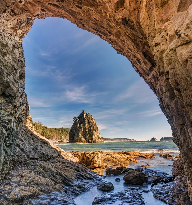 A rocky sea cave frames a view of a large, tree-topped sea stack rising from the blue ocean—a stunning scene along the Coastal PNW, with a rugged coastline and forested land under a partly cloudy sky. Perfect for Pacific Northwest day trips.