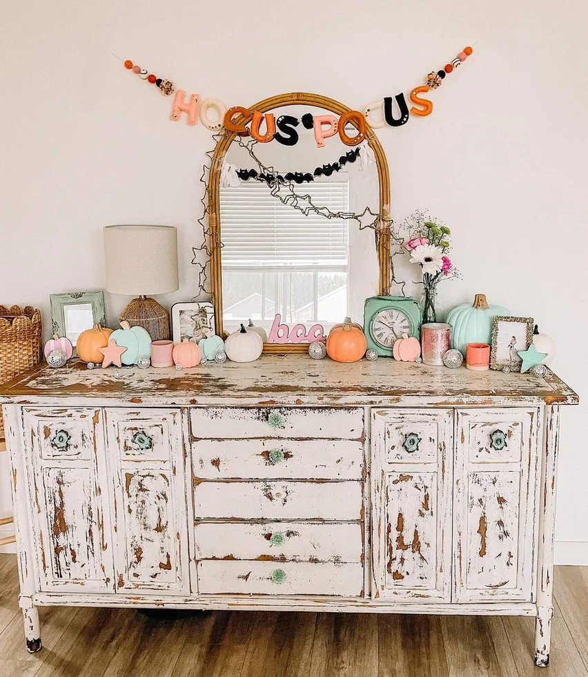 A rustic white dresser holds pastel pumpkins, candles, books, and decor. A mirror with string lights hangs above, with a HOCUS POCUS garland draped across it—perfect Halloween decor for creating a cozy, festive autumn vibe.