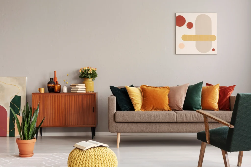 A modern living room with a brown sofa decorated with colorful cushions in warm, earthy colors inspired by Winter 2025 color trends, a wooden sideboard with vases and books, a potted plant, a yellow knit pouf, and abstract wall art on a light gray wall.