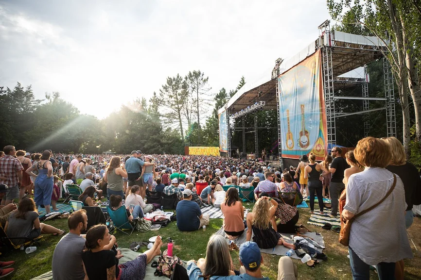 A large crowd sits and stands on a grassy area, enjoying a Concerts on the Lawn performance on an outdoor stage decorated with banners and surrounded by trees in the daylight.